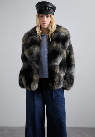 Γυναίκα που φοράει ένα μαύρο δερμάτινο καπέλο, γκρίζο και μαύρο παλτό από γούνα, ανοιχτό μπλε πουκάμισο με κουμπιά και φαρδύ σκούρο μπλε παντελόνι με σχέδια, όρθια μπροστά σε ουδέτερο φόντο.