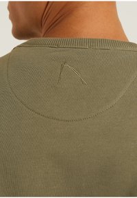 Gros plan sur le cou et le haut du dos d'une personne portant une chemise en maille vert olive avec des détails subtils cousus près du col.
