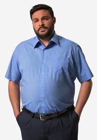 Camisa de manga corta de color azul claro con un acabado texturizado, con cuello puntiagudo, botonadura frontal y un bolsillo en el pecho. Llevada con pantalones oscuros.