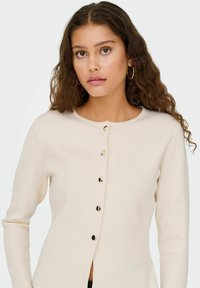 Cardigan en tricot beige avec un col rond, des manches longues et des fermetures à boutons dorés sur le devant. Texture lisse avec une silhouette ajustée.