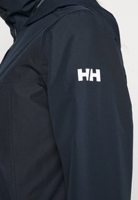 Laivastonsininen vedenpitävä takki, jossa on sileä pinta, etuvetoketju ja valkoinen logo vasemmassa hihassa. Lähikuva hihasta.