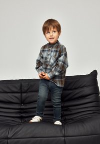 Karo dugačka košulja s dugim rukavima u plavoj, smeđoj i bijeloj boji; kopča se na gumbe, ima jedan džep na prsima. Kombinirana s tamnim trapericama i bijelim cipelama, sjedi na crnoj sofi.