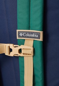 Cinghia dello zaino Columbia in verde e blu navale, con una resistente fibbia in plastica beige e un'etichetta logo cucita. Materiali in tessuto testurizzato.
