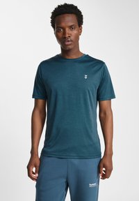 T-shirt de sport teal avec des manches courtes et un col rond. Comprend un petit logo blanc sur la poitrine gauche. Texture du tissu lisse.