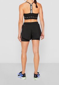 Mujer con sujetador deportivo negro de Puma, pantalones cortos de running negros en capas y zapatillas de running azules de Puma, de pie contra un fondo liso.