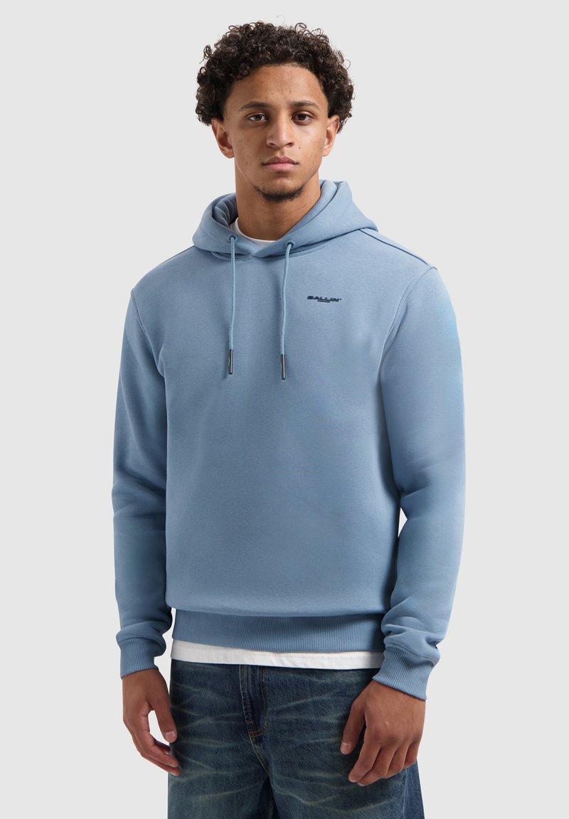 Jonge man met krullend haar, gekleed in een lichtblauwe hoodie over een wit shirt en blauwe jeans, staand tegen een eenvoudige lichte achtergrond.