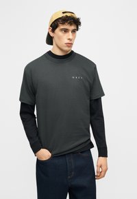 Νέος άνδρας που φοράει κίτρινο καπέλο με την κορυφή προς τα πίσω, σκούρο γκρι κοντομάνικη μπλούζα πάνω από μαύρο μακρυμάνικο πουκάμισο και σκούρο μπλε τζιν, κοιτάζοντας προς τα αριστερά.