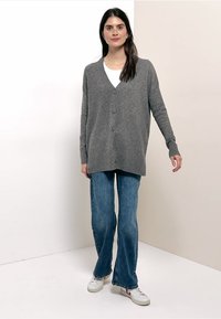 Grijze gebreide cardigan met een V-hals, vijf knopen en lange mouwen, gecombineerd met lichtblauwe uitlopende spijkerbroeken en witte sneakers.