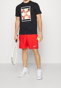 Camiseta gráfica negra para hombre con pelotas de tenis amarillas, combinada con pantalones cortos rojos y zapatillas blancas de Nike, sosteniendo una raqueta de tenis y una pelota.