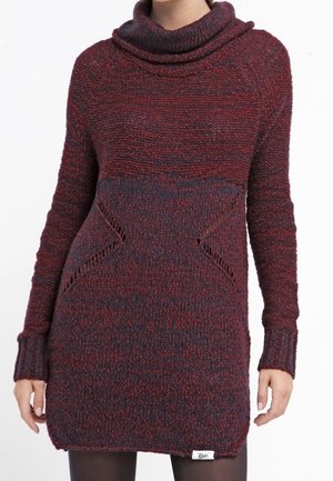 Vrouw draagt een lange, bordeauxrode en donkergrijze gebreide sweaterjurk met een colnek en textuurrijke driehoekige zijdetails.