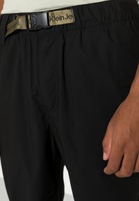 Pantaloni corti neri in elastico con un tessuto leggero e strutturato. Presentano una cintura beige con il logo "Klein Jeans" e tasche laterali su ciascun lato.
