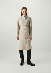 Vrouw met een glimlach, gekleed in een beige belted jas over zwarte cropped broeken en zwarte enkelboots, staande tegen een effen lichte achtergrond.