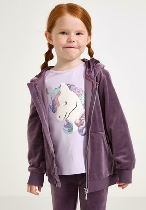 Jong meisje met rood haar en sproeten, gekleed in een paarse velours hoodie en broek, en een lavendel t-shirt met een pailletten eenhoorn ontwerp.