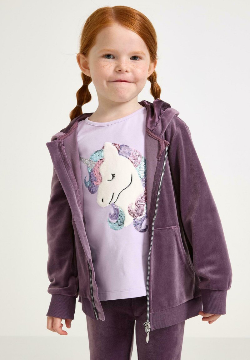 Ragazzina rossa con lentiggini che indossa un hoodie di velluto viola e pantaloni, e una maglietta lavanda con un design di unicorno in paillettes.