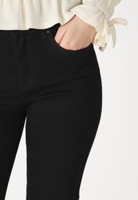 Personne portant un pantalon noir avec une main dans la poche avant et un haut à manches longues couleur crème avec des poignets noués.