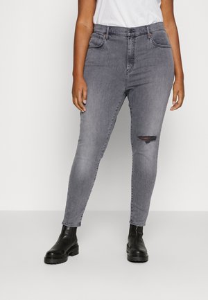 Person iført grå skinny jeans med en lille revne på højre lår, sorte ankelstøvler og en hvid indstoppet skjorte, stående mod en ensfarvet baggrund.