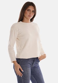 Maglione crema con scollatura rotonda, maniche lunghe e orlo a coste. Il tessuto ha una finitura strutturata, abbinato a jeans blu scuro.