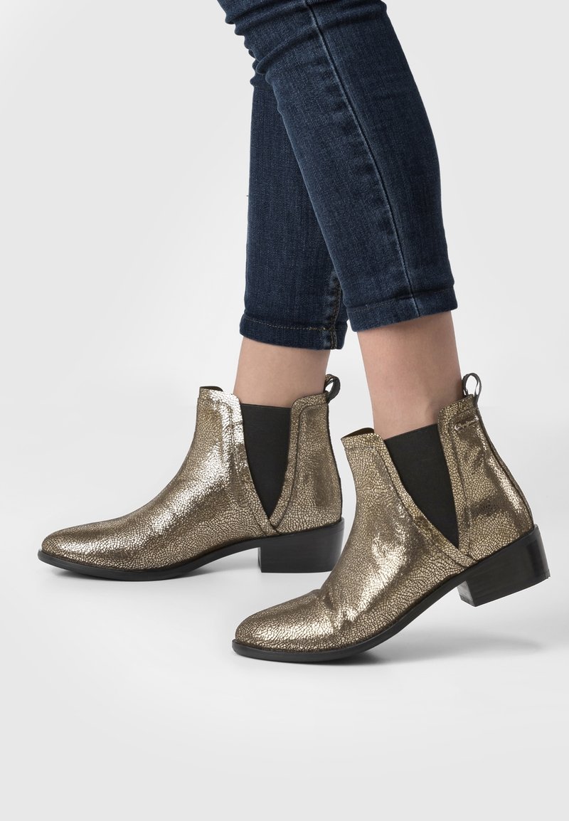 Personne portant des bottines métalliques dorées texturées avec des panels élastiques noirs sur les côtés et des talons bas noirs, associées à un jean skinny bleu foncé retroussé.