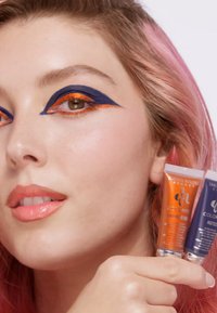 Dos tubos de cosméticos—uno naranja y otro azul profundo—sostenidos por una persona con cabello rosa, que lleva un maquillaje de ojos audaz en azul y naranja.