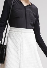 Schwarzes Langarmoberteil mit zwei Brusttaschen, kombiniert mit einem weißen strukturierten Rock im ausgestellten Design, gehalten von einem schwarzen Handtaschenriemen.