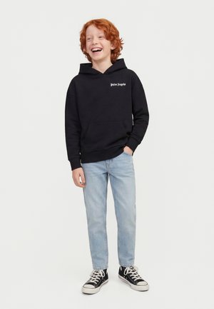 Chłopiec z rudymi włosami w czarnej bluzie z kapturem "Palm Angels", jasnoniebieskich dżinsach i czarnych wysokich trampkach, uśmiechający się z jedną ręką w kieszeni.