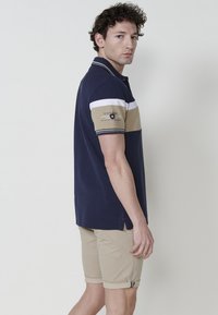 Sötétkék pólóing fehér és bézs vízszintes csíkokkal, rövid ujjú, és hímzett logóval a karon. Bézs feltekert sorttal viselve.