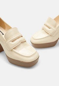 Mocassini in pelle verniciata beige con design a doppia tracolla, alta piattaforma e tacco in legno. Superficie liscia con punta arrotondata.