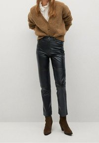 Pantalon en similicuir noir à coupe près du corps, avec des détails de coutures et une taille mi-haute, associé à un cardigan marron en tricot.