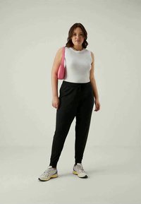 Zwarte joggingsbroek met een elastische tailleband, gecombineerd met een witte mouwloze top. Model draagt een kleine roze handtas en heeft multicolore sneakers aan.