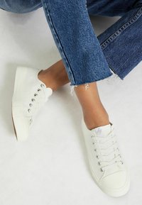 Baskets en toile blanches avec bout rond, semelle plate en caoutchouc et œillets en argent. Portées avec un jean en denim à jambe droite, avec un ourlet effiloché.