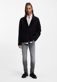 Blazer negro de gran tamaño con dos bolsillos delanteros, combinado con una camisa blanca, vaqueros ajustados grises y zapatos negros. Diseño sencillo, textura suave.