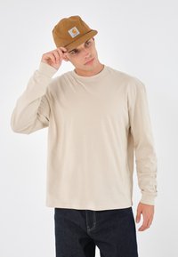 Beige langermet skjorte med rund hals, kombinert med en brun caps med et firkantet logo, og mørkeblå denim jeans.