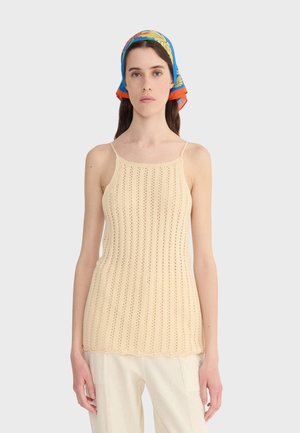 Donna che indossa un top smanicato lavorato a maglia color crema, pantaloni bianchi e un foulard colorato, in piedi contro uno sfondo chiaro uniforme.