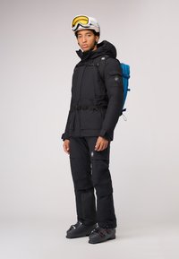 Zwarte geïsoleerde skijas met capuchon, cinched taille en verstelbare gespen. Gecombineerd met zwarte broek, skischoenen en een blauwe rugzak.