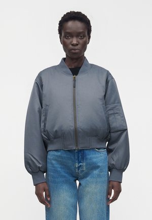 Femme portant une veste bomber grise zippée avec des poignets élastiques et un jean bleu, debout sur un fond blanc uni.