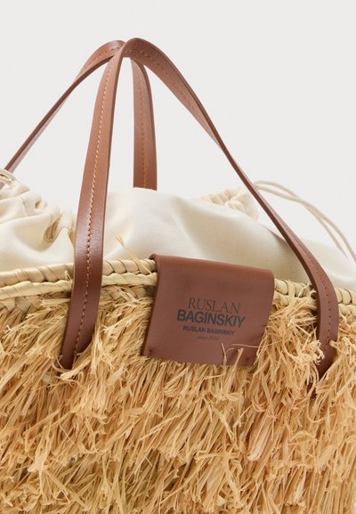 Τσάντα τύπου καλάθι από άχυρο με λαβές και ετικέτα από καφέ δέρμα, με κλείσιμο με κορδόνι από μπεζ ύφασμα στο εσωτερικό.