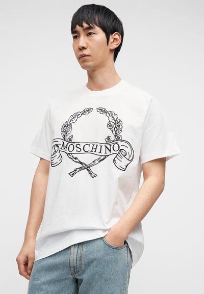 Λευκό βαμβακερό μπλουζάκι με μαύρο γραφικό σχέδιο με στεφάνι δάφνης και τη λέξη "MOSCHINO", συνδυασμένο με ανοιχτόχρωμο μπλε τζιν.