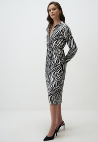 Zebraprint jurk met een kraag, lange mouwen, een taille met plooien en een knie-lange zoom. De stof lijkt zijdeachtig. Gekleed met zwarte hakken.