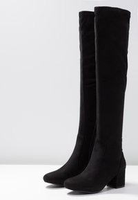 Botas negras de tacón alto, por encima de la rodilla, hechas de un suave material aterciopelado. Presentan una puntera redondeada y un talón de bloque robusto, con una silueta suave.