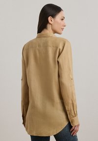 Majica z dolgimi rokavi iz svetlo rjavega, lanenega podobnega blaga. Ima ovratnik, gumbe na manšetah in zaobljen rob. Teksturiran zaključek.