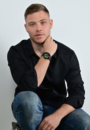 Black wristwatch with green accents, round face, and textured strap. Model wearing a black shirt and blue jeans, seated with hand on chin.