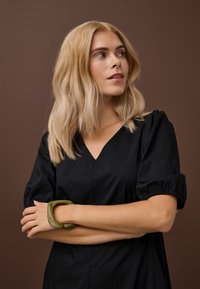 Plavolasa ženska v črni obleki z V-izrezom in napihnjenimi rokavi, sklenjenih rok, ki gleda vstran na rjavi podlagi.