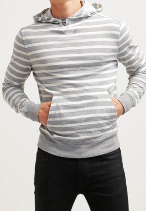 Homme portant un sweat à capuche rayé gris et blanc avec les mains dans la poche avant, associé à un pantalon noir, sur un fond uni.