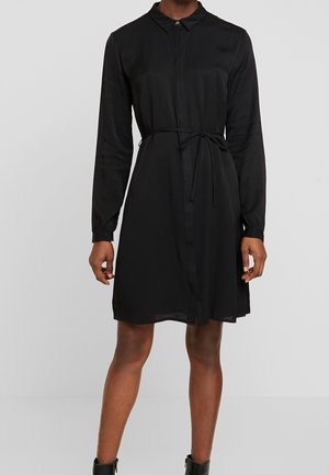 Femme portant une robe-chemise noire à manches longues avec un col et une ceinture à nouer, associée à des bottines noires, debout devant un fond uni.