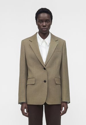 Personne noire portant un blazer beige à deux boutons sur une chemise blanche à col et un pantalon foncé, debout devant un fond clair uni.
