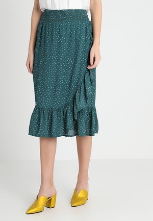 Jupe teal avec des pois blancs, taille smockée, ourlet asymétrique à volants. Portée avec des mules jaunes brillantes. Texture de tissu lisse.