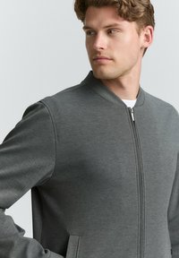 Szara kurtka typu bomber z wysokim kołnierzem, zapięciem na zamek i bocznymi kieszeniami; wykonana z gładkiego, miękkiego materiału o czystym, nowoczesnym designie.