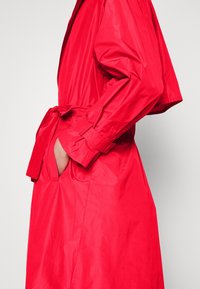 Czerwony wodoodporny płaszcz trench z pasem w talii, bocznymi kieszeniami i długimi rękawami. Charakteryzuje się gładką, lekką teksturą.