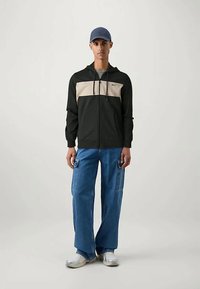 Mladý muž oblečený v čiernej a béžovej mikine s kapucňou, modrých cargo džínsoch, bielych teniskách a tmavomodrej šiltovke stojí proti jednoduchému svetlému pozadiu.