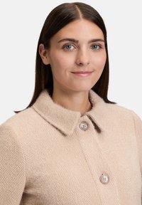 Młoda kobieta z prostymi ciemnymi włosami, nosząca beżową, teksturowaną wełnianą marynarkę z dużym kołnierzem i dwoma widocznymi guzikami.